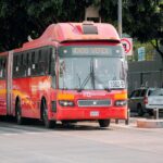 A red Metrobús navigating a city street surrounded by cars and greenery.