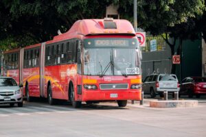 A red Metrobús navigating a city street surrounded by cars and greenery.